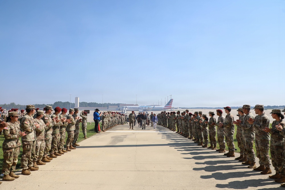 A formal military honor cordon ceremony is taking place on an airfield tarmac, with two long rows of soldiers in camouflage uniforms and red berets lining a walkway. A small group of individuals, including civilians and military personnel, walk between the rows while soldiers applaud. An American Airlines aircraft is visible in the background, suggesting this is a commercial or joint-use airport.