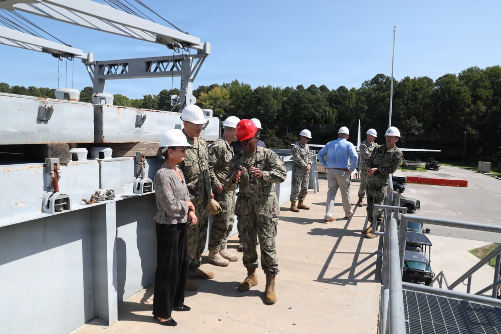 A group of U.S. Navy personnel in digital camouflage uniforms and civilians are conducting what appears to be an official tour or inspection of a naval facility or vessel. Most individuals are wearing white hard hats as required safety equipment, with one person wearing a red hard hat. The setting includes heavy industrial equipment such as a crane structure, metal decking, and a paved facility area surrounded by trees.