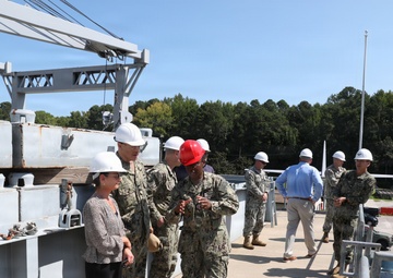 A group of U.S. Navy personnel in digital camouflage uniforms and civilians are conducting what appears to be an official tour or inspection of a naval facility or vessel. Most individuals are wearing white hard hats as required safety equipment, with one person wearing a red hard hat. The setting includes heavy industrial equipment such as a crane structure, metal decking, and a paved facility area surrounded by trees.