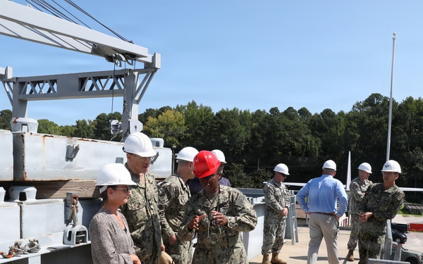 A group of U.S. Navy personnel in digital camouflage uniforms and civilians are conducting what appears to be an official tour or inspection of a naval facility or vessel. Most individuals are wearing white hard hats as required safety equipment, with one person wearing a red hard hat. The setting includes heavy industrial equipment such as a crane structure, metal decking, and a paved facility area surrounded by trees.