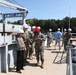 A group of U.S. Navy personnel in digital camouflage uniforms and civilians are conducting what appears to be an official tour or inspection of a naval facility or vessel. Most individuals are wearing white hard hats as required safety equipment, with one person wearing a red hard hat. The setting includes heavy industrial equipment such as a crane structure, metal decking, and a paved facility area surrounded by trees.