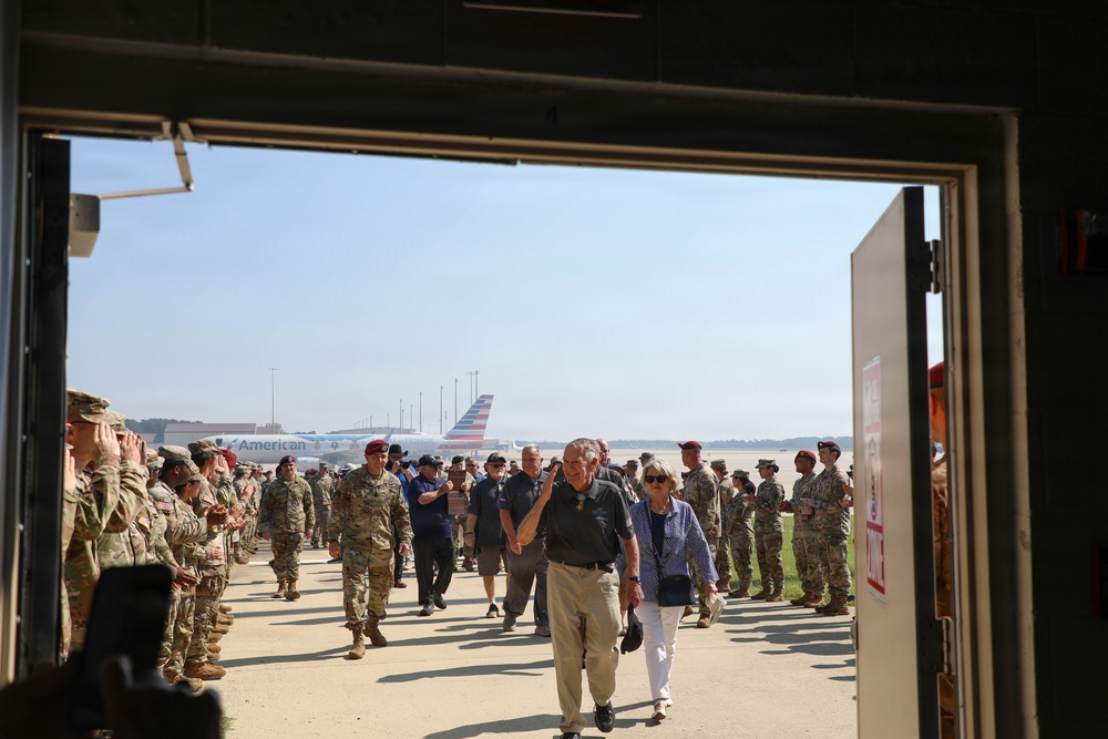 A military ceremony or welcome event is taking place on an airfield tarmac, viewed through a large hangar doorway. Uniformed military personnel in camouflage uniforms are lined up on both sides forming an honor corridor, while civilians walk through the middle toward the hangar entrance. An American Airlines aircraft is visible in the background on the tarmac.