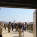 A military ceremony or welcome event is taking place on an airfield tarmac, viewed through a large hangar doorway. Uniformed military personnel in camouflage uniforms are lined up on both sides forming an honor corridor, while civilians walk through the middle toward the hangar entrance. An American Airlines aircraft is visible in the background on the tarmac.