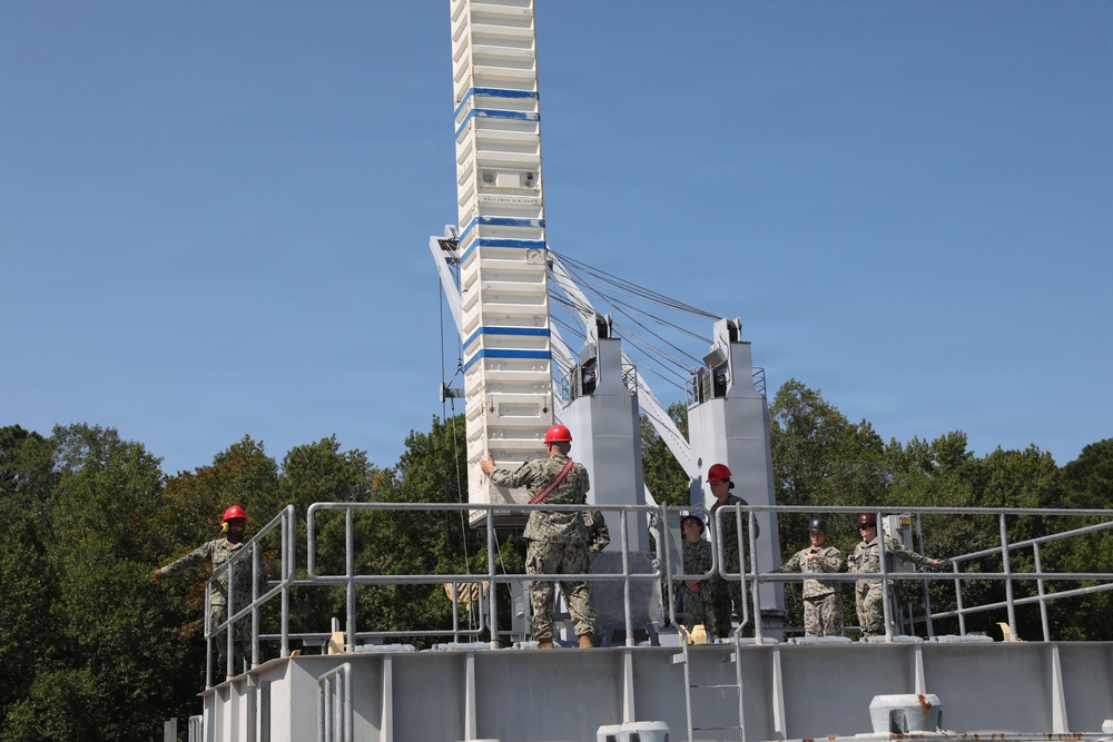 Military personnel in camouflage uniforms are gathered on an elevated platform or deck of what appears to be a naval training facility or vessel simulator. Several individuals are wearing red hard hats, indicating a safety-regulated work or training environment. A large white tower structure with cables and mechanical components dominates the background.