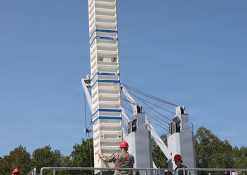 Military personnel in camouflage uniforms are gathered on an elevated platform or deck of what appears to be a naval training facility or vessel simulator. Several individuals are wearing red hard hats, indicating a safety-regulated work or training environment. A large white tower structure with cables and mechanical components dominates the background.