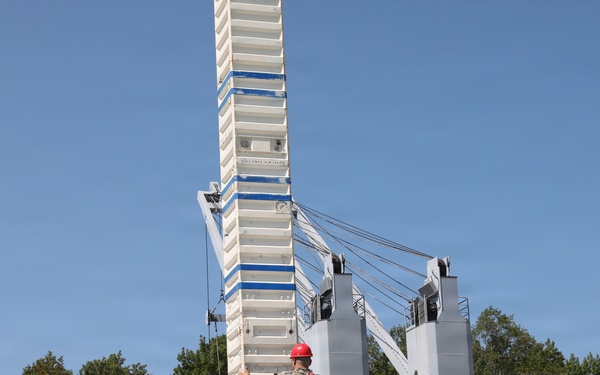 Military personnel in camouflage uniforms are gathered on an elevated platform or deck of what appears to be a naval training facility or vessel simulator. Several individuals are wearing red hard hats, indicating a safety-regulated work or training environment. A large white tower structure with cables and mechanical components dominates the background.