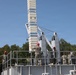 Military personnel in camouflage uniforms are gathered on an elevated platform or deck of what appears to be a naval training facility or vessel simulator. Several individuals are wearing red hard hats, indicating a safety-regulated work or training environment. A large white tower structure with cables and mechanical components dominates the background.