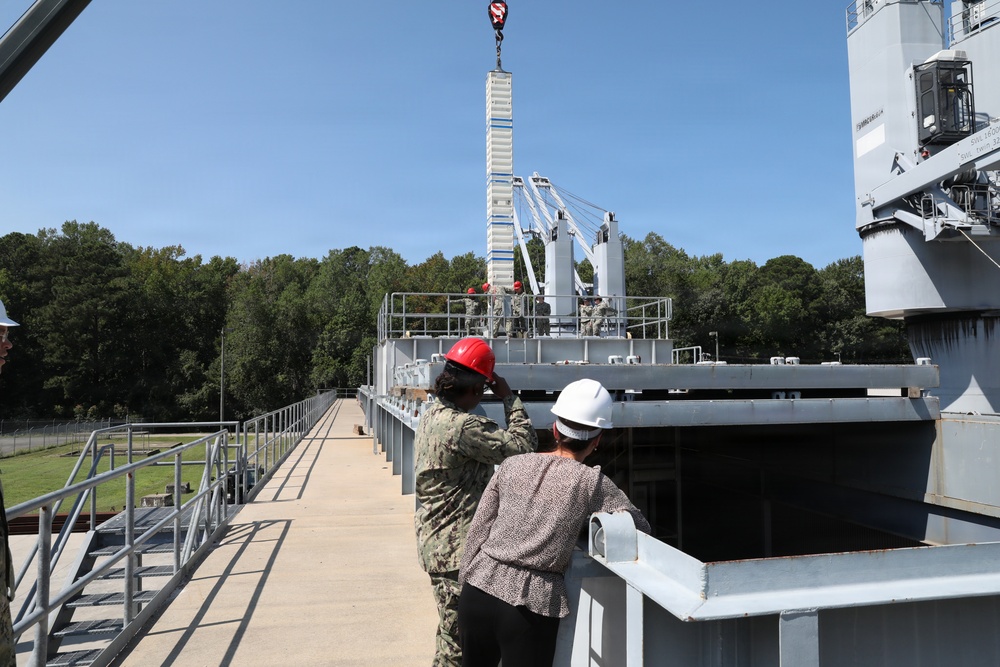 Two individuals wearing hard hats — one in a military uniform with a red hard hat and one in civilian attire with a white hard hat — observe a crane lifting operations at what appears to be a naval or military industrial facility. Workers can be seen on an elevated platform in the background overseeing the lift. The setting includes metal walkways, railings, and large gray industrial structures surrounded by trees.
