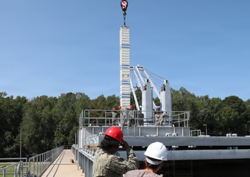 Two individuals wearing hard hats — one in a military uniform with a red hard hat and one in civilian attire with a white hard hat — observe a crane lifting operations at what appears to be a naval or military industrial facility. Workers can be seen on an elevated platform in the background overseeing the lift. The setting includes metal walkways, railings, and large gray industrial structures surrounded by trees.