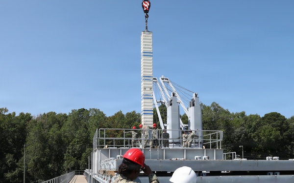 Two individuals wearing hard hats — one in a military uniform with a red hard hat and one in civilian attire with a white hard hat — observe a crane lifting operations at what appears to be a naval or military industrial facility. Workers can be seen on an elevated platform in the background overseeing the lift. The setting includes metal walkways, railings, and large gray industrial structures surrounded by trees.