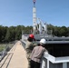 Two individuals wearing hard hats — one in a military uniform with a red hard hat and one in civilian attire with a white hard hat — observe a crane lifting operations at what appears to be a naval or military industrial facility. Workers can be seen on an elevated platform in the background overseeing the lift. The setting includes metal walkways, railings, and large gray industrial structures surrounded by trees.