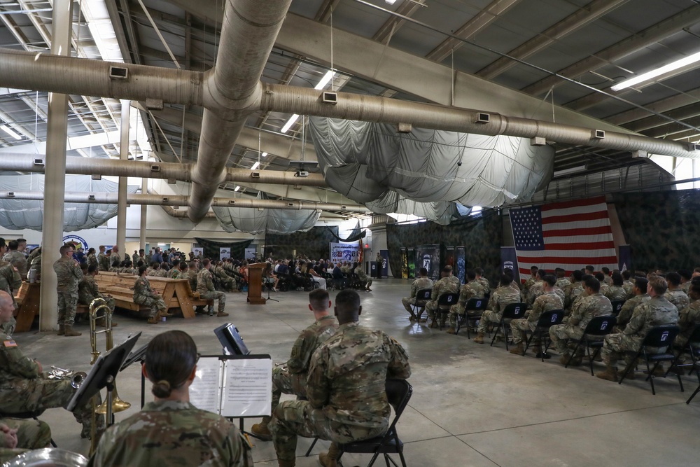 A formal military ceremony is taking place inside a large hangar or warehouse facility. Numerous soldiers in Army Combat Uniforms (ACU/OCP pattern) are seated in rows of chairs facing a podium and stage area decorated with an American flag and camouflage netting. A military band is visible in the foreground, with musicians seated at music stands holding brass instruments.