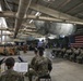 A formal military ceremony is taking place inside a large hangar or warehouse facility. Numerous soldiers in Army Combat Uniforms (ACU/OCP pattern) are seated in rows of chairs facing a podium and stage area decorated with an American flag and camouflage netting. A military band is visible in the foreground, with musicians seated at music stands holding brass instruments.