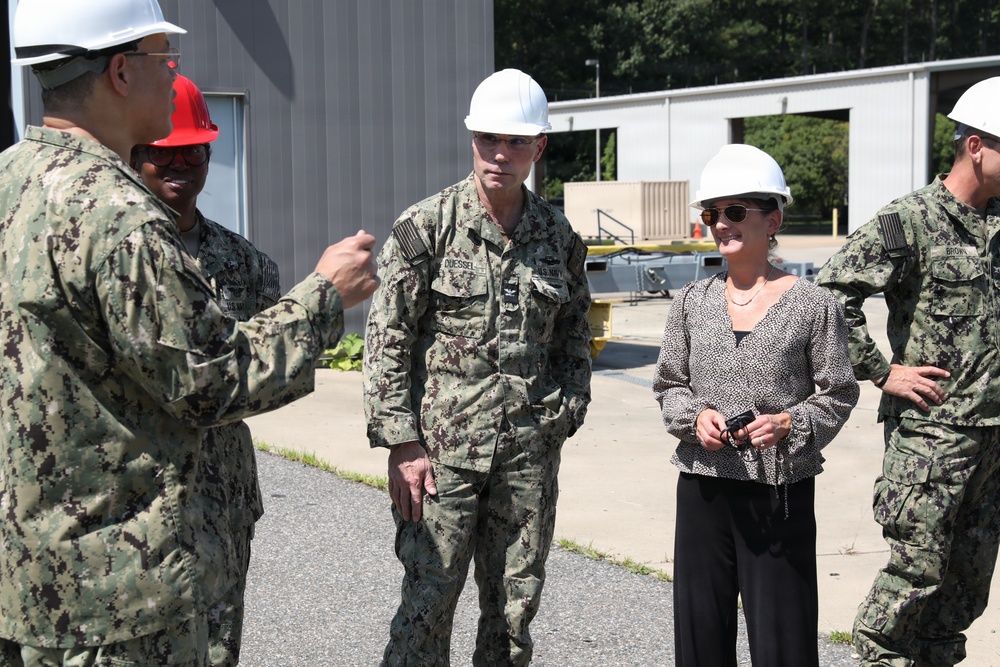 A group of U.S. Navy personnel in NWU Type III camouflage uniforms and a civilian woman are gathered outdoors at what appears to be a military installation or facility. All individuals are wearing hard hats, suggesting a safety-required area such as a construction or industrial site tour. One uniformed individual appears to be briefing or explaining something to the group.