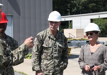 A group of U.S. Navy personnel in NWU Type III camouflage uniforms and a civilian woman are gathered outdoors at what appears to be a military installation or facility. All individuals are wearing hard hats, suggesting a safety-required area such as a construction or industrial site tour. One uniformed individual appears to be briefing or explaining something to the group.