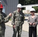 A group of U.S. Navy personnel in NWU Type III camouflage uniforms and a civilian woman are gathered outdoors at what appears to be a military installation or facility. All individuals are wearing hard hats, suggesting a safety-required area such as a construction or industrial site tour. One uniformed individual appears to be briefing or explaining something to the group.