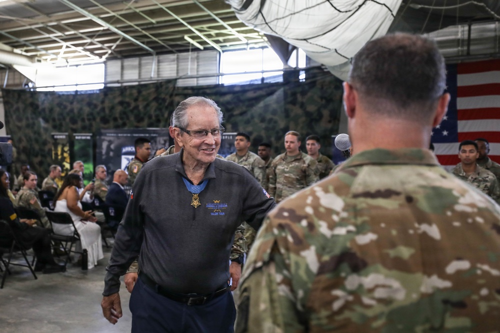 An elderly civilian man wearing a Medal of Honor around his neck is engaged in conversation with a uniformed military soldier during what appears to be a formal military ceremony or event. The setting is an indoor military facility, likely a hangar or large bay, decorated with camouflage netting and an American flag. A crowd of soldiers and civilians are seated and standing in the background.
