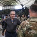 An elderly civilian man wearing a Medal of Honor around his neck is engaged in conversation with a uniformed military soldier during what appears to be a formal military ceremony or event. The setting is an indoor military facility, likely a hangar or large bay, decorated with camouflage netting and an American flag. A crowd of soldiers and civilians are seated and standing in the background.