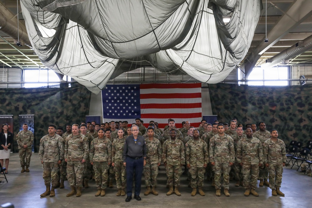 A large group of U.S. Army soldiers in camouflage uniforms are assembled in formation inside a military hangar or large indoor facility. They are posed for a group photograph in front of a large American flag mounted on a camouflage-patterned backdrop. One civilian individual stands at the center front of the formation, distinguished from the uniformed personnel.