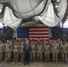A large group of U.S. Army soldiers in camouflage uniforms are assembled in formation inside a military hangar or large indoor facility. They are posed for a group photograph in front of a large American flag mounted on a camouflage-patterned backdrop. One civilian individual stands at the center front of the formation, distinguished from the uniformed personnel.
