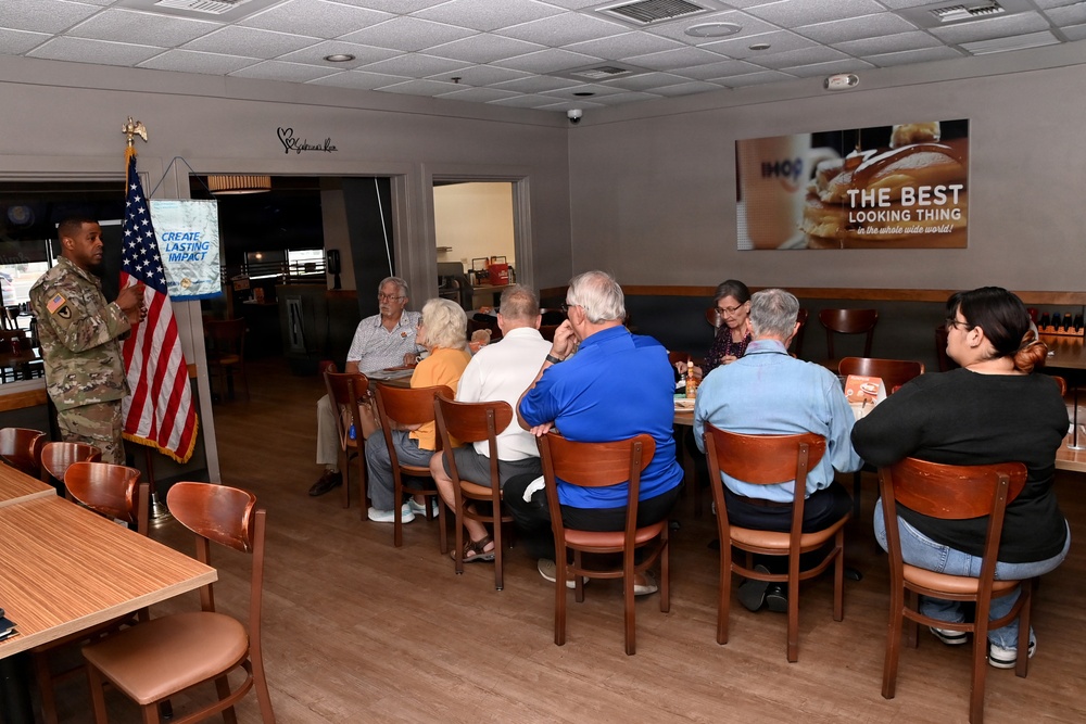 A U.S. Army soldier in camouflage uniform is addressing a small group of civilians seated at tables in an IHOP restaurant. A banner reading 'Create Lasting Impact' and an American flag are positioned beside the speaker. The gathering appears to be a community or civic meeting, possibly a Rotary Club or similar organization event.