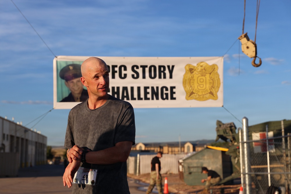 A bald man wearing a gray t-shirt and a race bib (number 49) stands in the foreground at what appears to be a military fitness challenge event. Behind him is a banner reading 'PFC STORY CHALLENGE' featuring a military portrait photo and a unit insignia with the number 9. The setting appears to be a military installation or base, with military equipment, personnel, and infrastructure visible in the background.