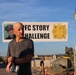 A bald man wearing a gray t-shirt and a race bib (number 49) stands in the foreground at what appears to be a military fitness challenge event. Behind him is a banner reading 'PFC STORY CHALLENGE' featuring a military portrait photo and a unit insignia with the number 9. The setting appears to be a military installation or base, with military equipment, personnel, and infrastructure visible in the background.