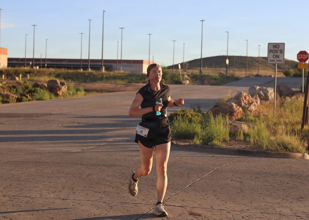 A female runner wearing bib number 48 is participating in a road race on a paved surface in what appears to be a desert or arid environment. She is dressed in black athletic wear and is carrying a small water bottle in her left hand. The setting appears to be a military installation or restricted area, indicated by signage reading 'NO POV'S ON TANK TRAIL' and a stop sign.