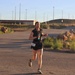 A female runner wearing bib number 48 is participating in a road race on a paved surface in what appears to be a desert or arid environment. She is dressed in black athletic wear and is carrying a small water bottle in her left hand. The setting appears to be a military installation or restricted area, indicated by signage reading 'NO POV'S ON TANK TRAIL' and a stop sign.