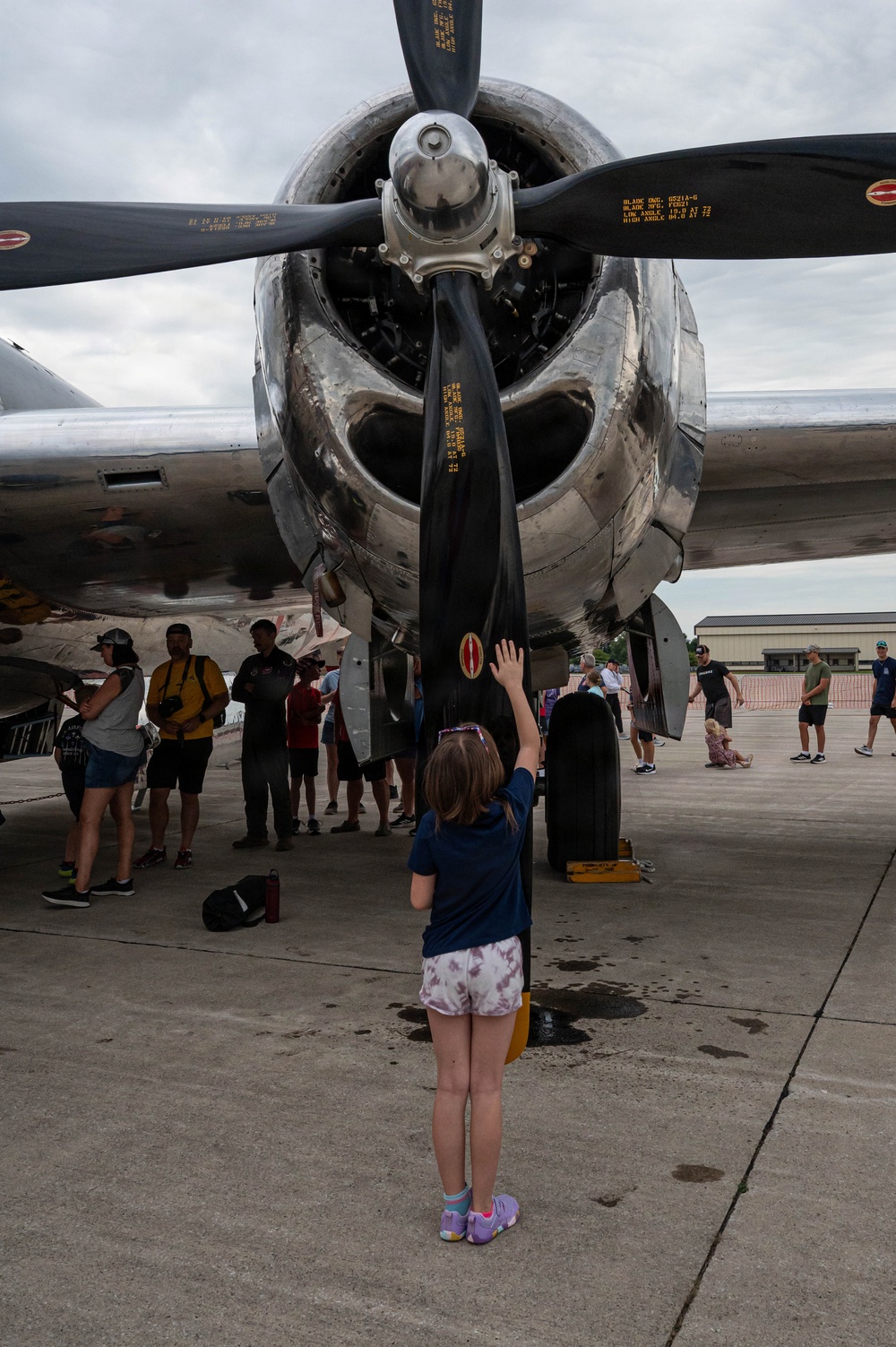 A young girl stands beneath a large radial engine and propeller of a vintage WWII-era bomber aircraft, reaching up to touch one of the propeller blades. The scene appears to be an airshow or static aircraft display event, with numerous spectators visible in the background examining the aircraft. The polished aluminum engine cowling and black propeller blades dominate the foreground.