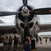 A young girl stands beneath a large radial engine and propeller of a vintage WWII-era bomber aircraft, reaching up to touch one of the propeller blades. The scene appears to be an airshow or static aircraft display event, with numerous spectators visible in the background examining the aircraft. The polished aluminum engine cowling and black propeller blades dominate the foreground.