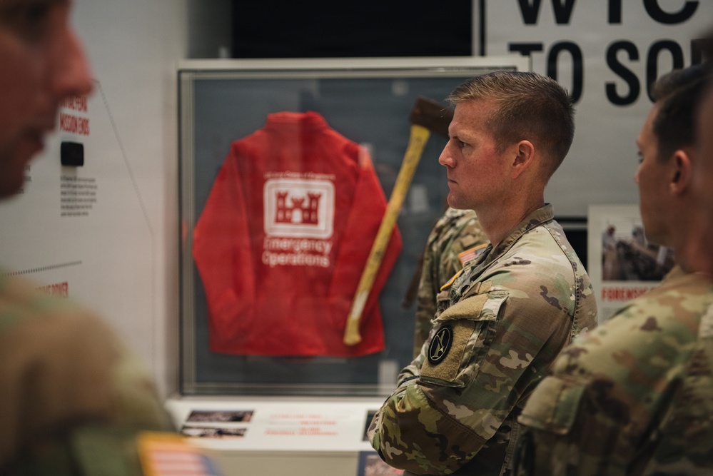 Military personnel in camouflage uniforms are viewing a museum or exhibit display. The central display case contains a red jacket labeled 'Emergency Operations' bearing a U.S. Army Corps of Engineers castle insignia, alongside what appears to be an axe or tool. The soldiers appear to be on a guided tour or educational visit.