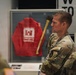 Military personnel in camouflage uniforms are viewing a museum or exhibit display. The central display case contains a red jacket labeled 'Emergency Operations' bearing a U.S. Army Corps of Engineers castle insignia, alongside what appears to be an axe or tool. The soldiers appear to be on a guided tour or educational visit.