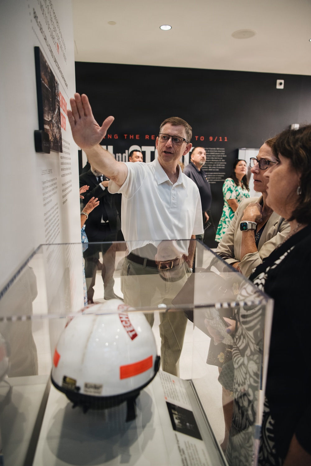A group of visitors are touring what appears to be a museum exhibition related to 9/11, as indicated by visible wall text reading 'THE RE... TO 9/11'. A man in a white polo shirt is gesturing toward wall-mounted displays while speaking to attendees, appearing to serve as a guide or presenter. In the foreground, a white spherical object with orange and red markings is displayed in a glass case.