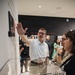 A group of visitors are touring what appears to be a museum exhibition related to 9/11, as indicated by visible wall text reading 'THE RE... TO 9/11'. A man in a white polo shirt is gesturing toward wall-mounted displays while speaking to attendees, appearing to serve as a guide or presenter. In the foreground, a white spherical object with orange and red markings is displayed in a glass case.