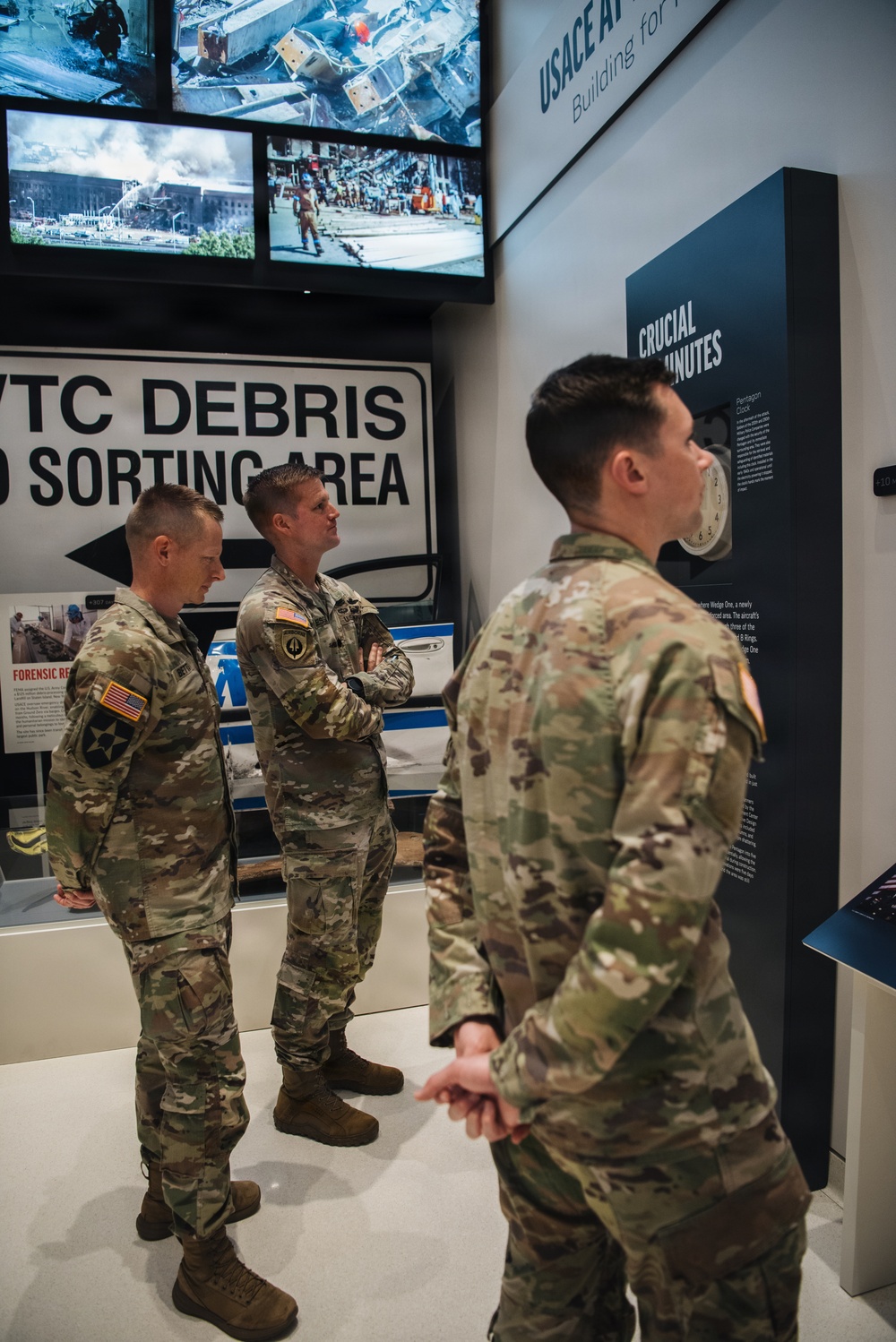 Three U.S. Army soldiers in OCP (Operational Camouflage Pattern) uniforms are touring what appears to be a 9/11 memorial museum exhibit. The exhibit features a 'WTC Debris Sorting Area' sign, aerial and ground-level photographs of the World Trade Center and Pentagon attack sites, and a display panel labeled 'Crucial Minutes' referencing a Pentagon Clock. The soldiers appear to be engaged in conversation while viewing the exhibits.