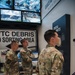 Three U.S. Army soldiers in OCP (Operational Camouflage Pattern) uniforms are touring what appears to be a 9/11 memorial museum exhibit. The exhibit features a 'WTC Debris Sorting Area' sign, aerial and ground-level photographs of the World Trade Center and Pentagon attack sites, and a display panel labeled 'Crucial Minutes' referencing a Pentagon Clock. The soldiers appear to be engaged in conversation while viewing the exhibits.