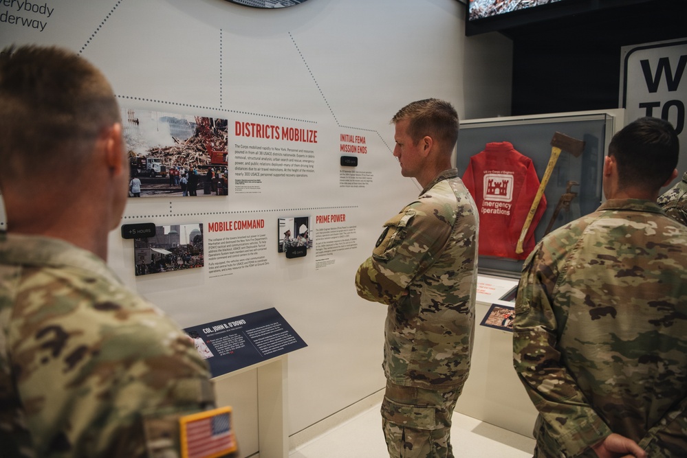 Military personnel in U.S. Army camouflage uniforms are viewing a museum exhibit about disaster response operations. The exhibit features sections titled 'Districts Mobilize,' 'Mobile Command,' 'Prime Power,' and 'Initial FEMA Mission Ends,' with photographs and artifacts related to emergency operations. A red 'Emergency Operations' jacket and an axe are displayed in a case on the right side of the exhibit.