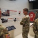 Military personnel in U.S. Army camouflage uniforms are viewing a museum exhibit about disaster response operations. The exhibit features sections titled 'Districts Mobilize,' 'Mobile Command,' 'Prime Power,' and 'Initial FEMA Mission Ends,' with photographs and artifacts related to emergency operations. A red 'Emergency Operations' jacket and an axe are displayed in a case on the right side of the exhibit.