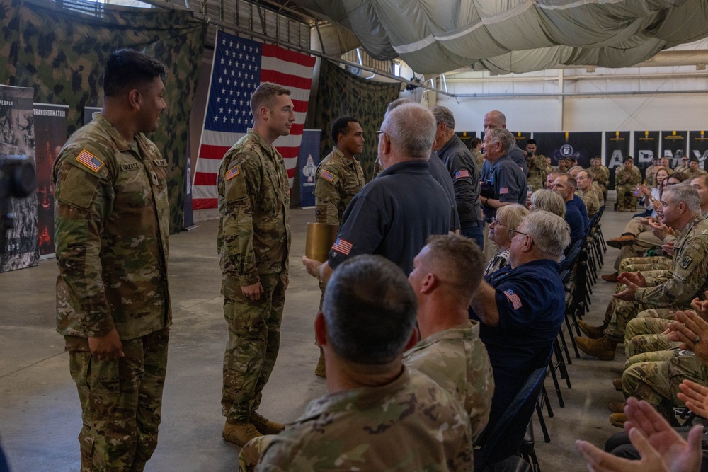 A military ceremony is taking place inside a large indoor facility, likely a hangar or armory, decorated with American flags and camouflage netting. Several soldiers in Army Combat Uniforms (ACU) are standing at attention while an audience of both military personnel and civilians applauds. The setting suggests an awards ceremony, promotion, or recognition event.