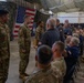 A military ceremony is taking place inside a large indoor facility, likely a hangar or armory, decorated with American flags and camouflage netting. Several soldiers in Army Combat Uniforms (ACU) are standing at attention while an audience of both military personnel and civilians applauds. The setting suggests an awards ceremony, promotion, or recognition event.