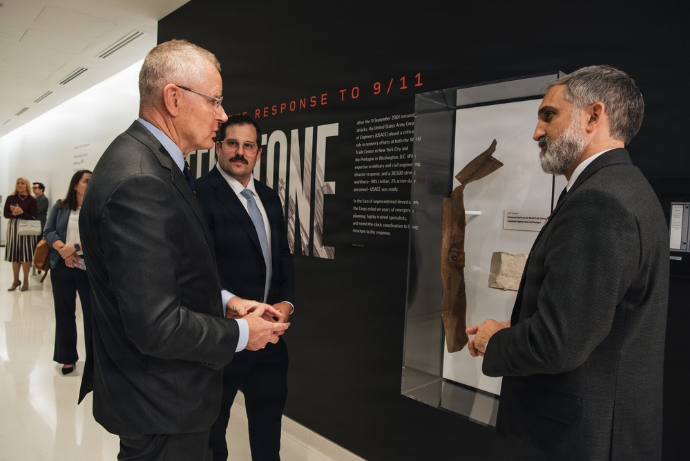 Three men in business attire are engaged in conversation at what appears to be a museum or memorial exhibition focused on '9/11 Response.' A display case containing artifacts, including what appears to be structural debris, is visible beside them. The exhibit wall features text about the United States Army Corps of Engineers' role in the September 11, 2001 terrorist attack response.