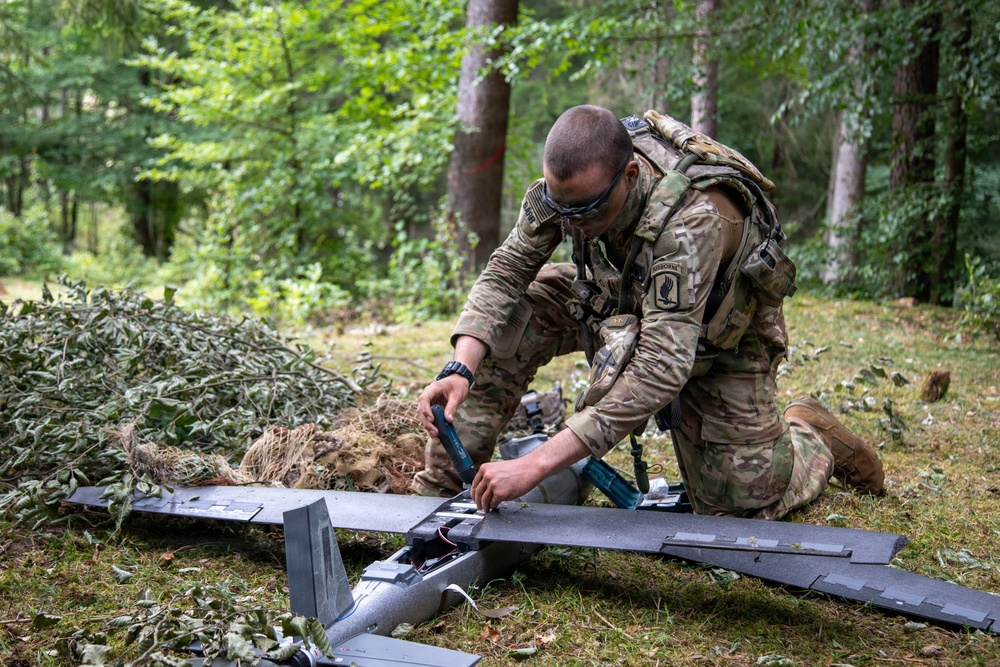 A U.S. Army soldier wearing Airborne unit patches and MultiCam camouflage is kneeling on the ground in a forested area, assembling or maintaining a fixed-wing unmanned aerial vehicle (UAV). The soldier is equipped with tactical gear including a backpack and protective eyewear. The setting appears to be a field training exercise or operational deployment in a woodland environment.