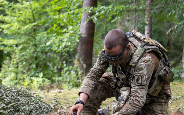 A U.S. Army soldier wearing Airborne unit patches and MultiCam camouflage is kneeling on the ground in a forested area, assembling or maintaining a fixed-wing unmanned aerial vehicle (UAV). The soldier is equipped with tactical gear including a backpack and protective eyewear. The setting appears to be a field training exercise or operational deployment in a woodland environment.