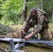 A U.S. Army soldier wearing Airborne unit patches and MultiCam camouflage is kneeling on the ground in a forested area, assembling or maintaining a fixed-wing unmanned aerial vehicle (UAV). The soldier is equipped with tactical gear including a backpack and protective eyewear. The setting appears to be a field training exercise or operational deployment in a woodland environment.