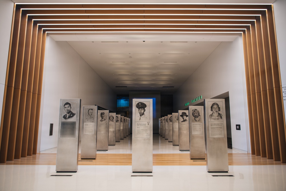 A modern museum gallery features rows of metallic memorial pedestals/stanchions displaying portrait illustrations and biographical text of individuals. The space is architecturally striking with concentric wooden frame elements and integrated LED lighting creating a tunnel-like entrance. A blue illuminated display screen is visible at the far end of the corridor.