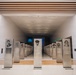A modern museum gallery features rows of metallic memorial pedestals/stanchions displaying portrait illustrations and biographical text of individuals. The space is architecturally striking with concentric wooden frame elements and integrated LED lighting creating a tunnel-like entrance. A blue illuminated display screen is visible at the far end of the corridor.
