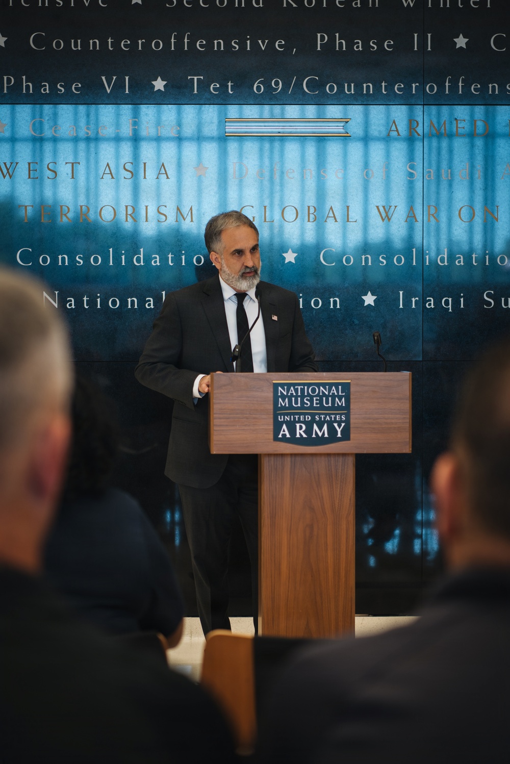 A man in a dark suit with a black tie and American flag lapel pin is speaking at a wooden podium bearing the 'National Museum United States Army' logo. The background displays a large panel with military campaign names and operations including 'Global War on Terrorism,' 'West Asia,' 'Cease-Fire,' and others. An audience is visible in the foreground, seen from behind.