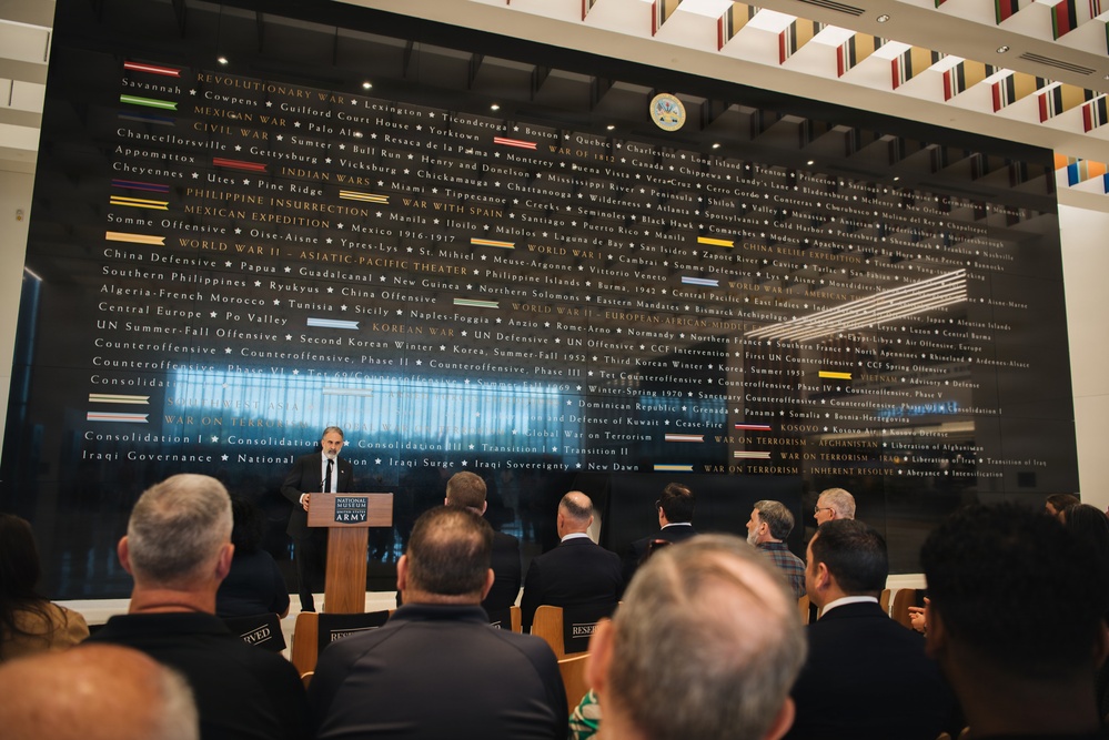 A speaker addresses an audience at a podium bearing the National Museum of the United States Army seal, set against a large black wall displaying the history of U.S. Army campaigns from the Revolutionary War through modern conflicts. The wall lists battles, campaigns, and military operations chronologically with colored unit citation ribbons beside each era. The setting appears to be a formal ceremony or dedication event at the museum.