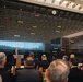 A speaker addresses an audience at a podium bearing the National Museum of the United States Army seal, set against a large black wall displaying the history of U.S. Army campaigns from the Revolutionary War through modern conflicts. The wall lists battles, campaigns, and military operations chronologically with colored unit citation ribbons beside each era. The setting appears to be a formal ceremony or dedication event at the museum.
